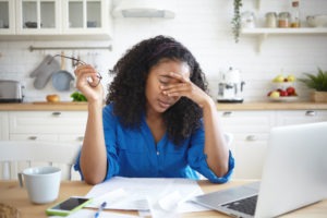 Sexual Harassment Lawsuit Loans | Get Up to $1 Million An African-American woman holding glasses sits at a kitchen table with bills and a laptop, stressed over her finances.