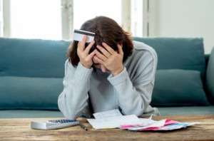 Traumatic Brain Injury Legal Funding | No Credit Check Required A young woman looks worried as she holds her head and a credit card in her hands in front of a coffee table full of bills.